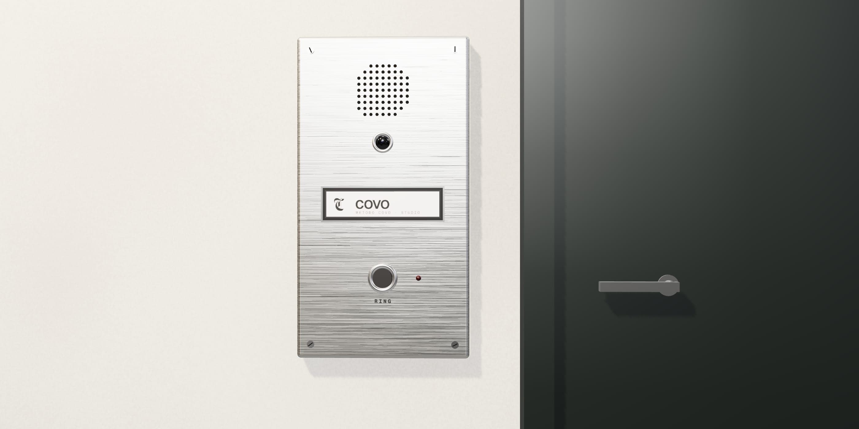 An intercom panel in brushed steel on a plaster wall: a speaker grille, a camera eye, a nameplate reading COVO, and a button marked RING, with a door beside it.